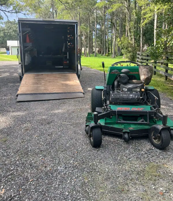Stand-on mower parked next to an open enclosed trailer with racked tools