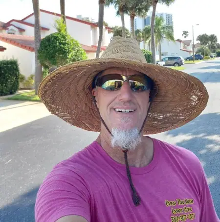 Over The Moon Lawn Care owner in a straw hat and company shirt on a Florida street lined with palms